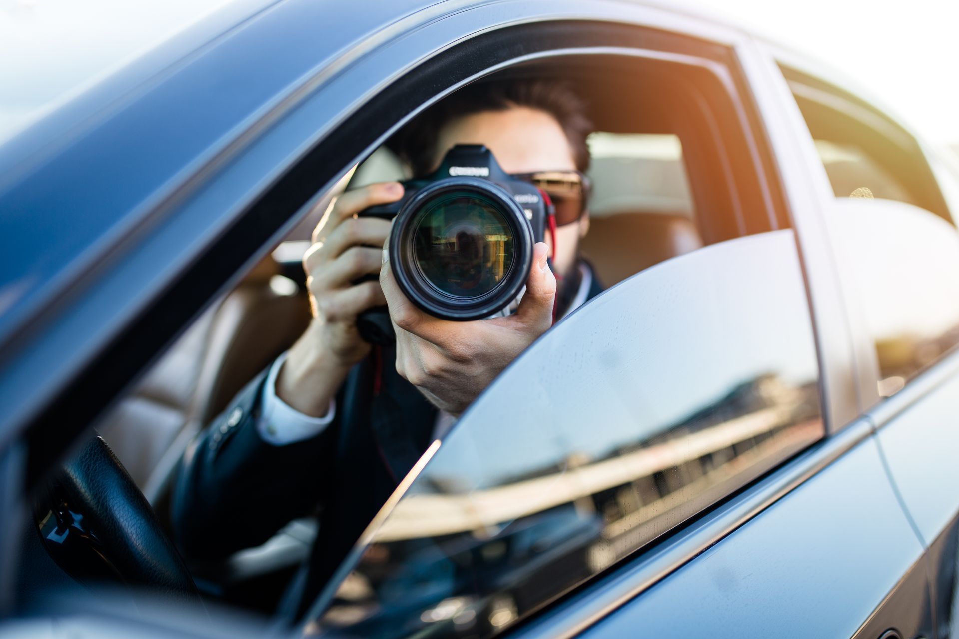 Photographer aiming a camera out of a car window, with warm sunlight reflecting on the blue car.