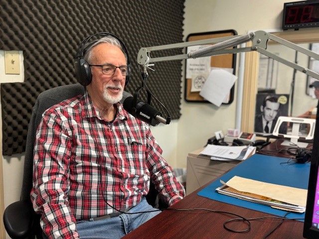 Man in plaid shirt wearing headphones, speaking into a microphone in a radio studio.