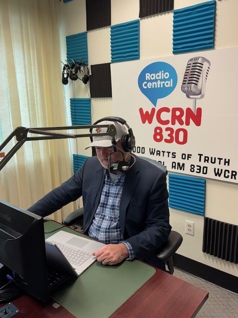 Man in radio studio at WCRN 830, reading papers, wearing headset and cap, blue and black soundproofing on wall.