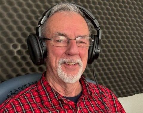 Older man with glasses and headphones, smiling, wearing a red plaid shirt, in front of soundproofing foam.