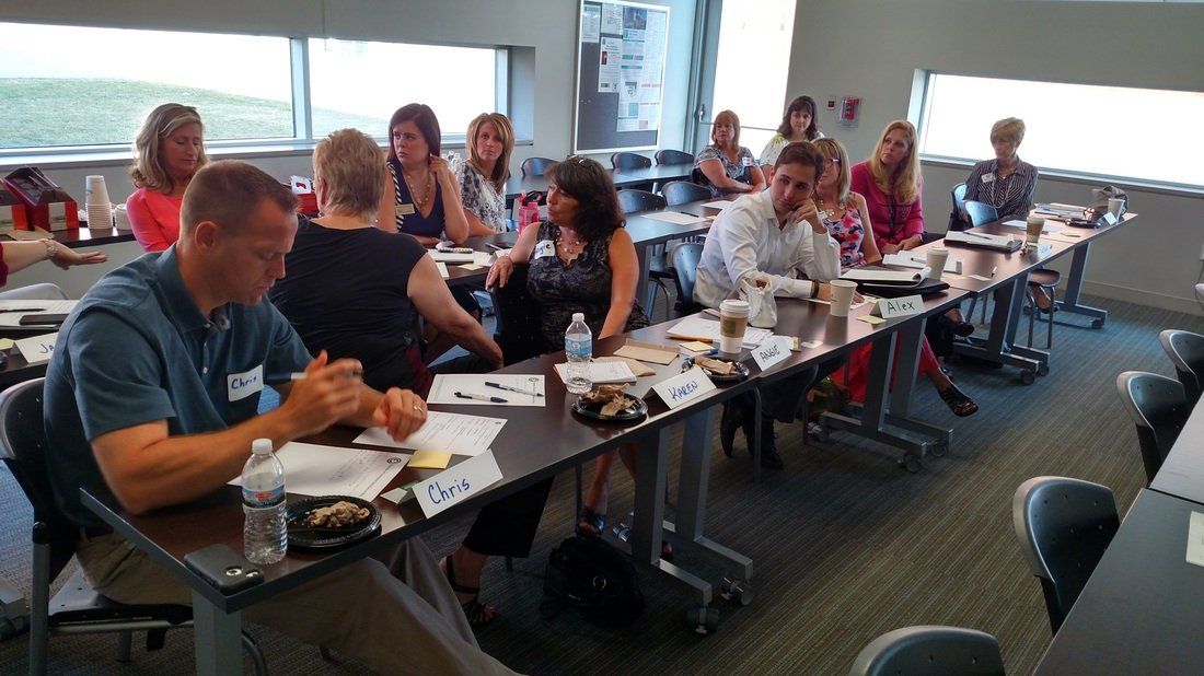 A group of people are sitting at long tables in a conference room.