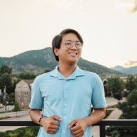 Man in blue shirt, smiling, wearing glasses, standing on a balcony with mountain backdrop.