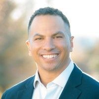 Man in a blue suit smiling at the camera, outdoors with blurred background.