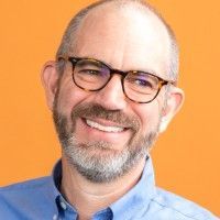 Man with glasses, smiling, blue shirt, against an orange background.