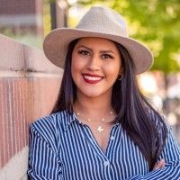 Woman in striped shirt and hat smiles, leans on a brick wall.