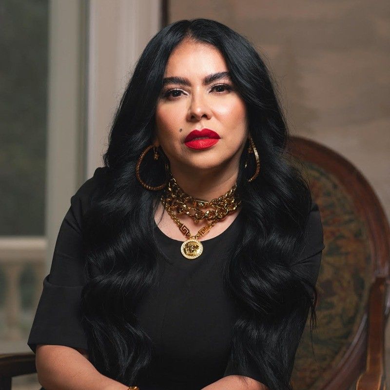 Woman with dark hair, red lipstick, and gold jewelry, wearing a black top, seated.