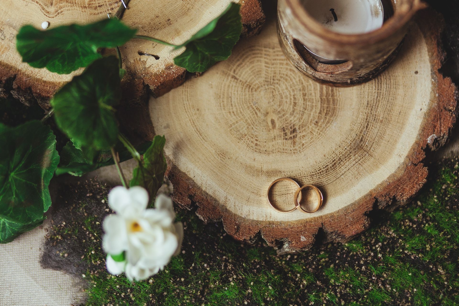 Due fedi nuziali sono appoggiate su una fetta di legno, accanto a un fiore e a un portacandele.