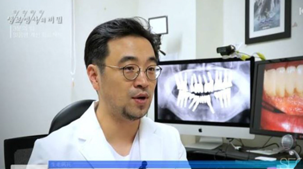 A dentist in a white coat speaks to the camera in his office, with dental x-rays and a close-up of teeth on computer screens behind him.