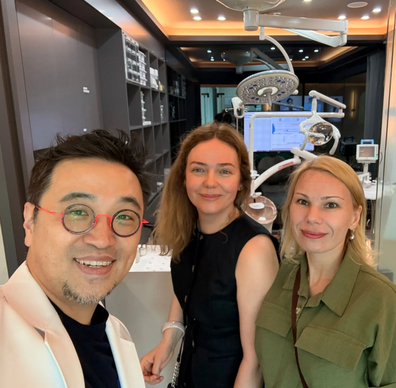 A woman and a dentist pose together in a dental exam room. The woman smiles while sitting in a dental chair, and the dentist smiles, wearing a mask.