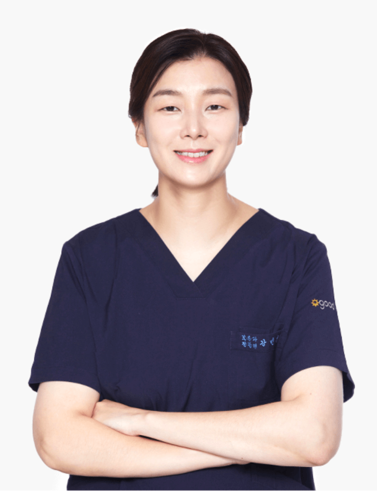 A smiling woman in dark blue scrubs with arms crossed, possibly a medical professional. Plain, neutral background.