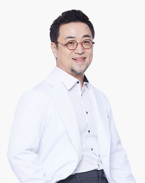 Man in a white lab coat smiling, wearing glasses. He has dark hair and a slight beard, set against a white background.