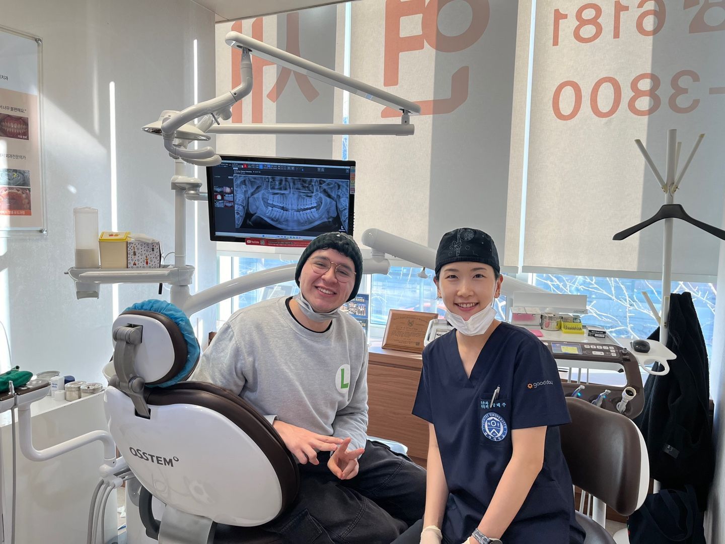 A smiling man and a dentist in a dental office. The dentist wears scrubs and a cap. The man sits in the dentist chair, both smiling.