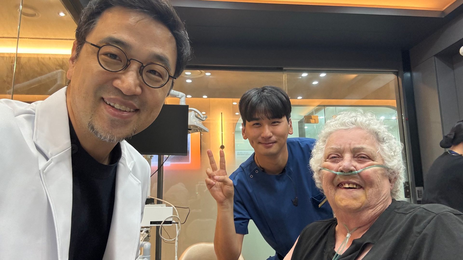 Three people smiling for a selfie in a dental clinic. An elderly woman with an oxygen tube sits, a male assistant makes a peace sign, and a doctor in a lab coat takes the photo.