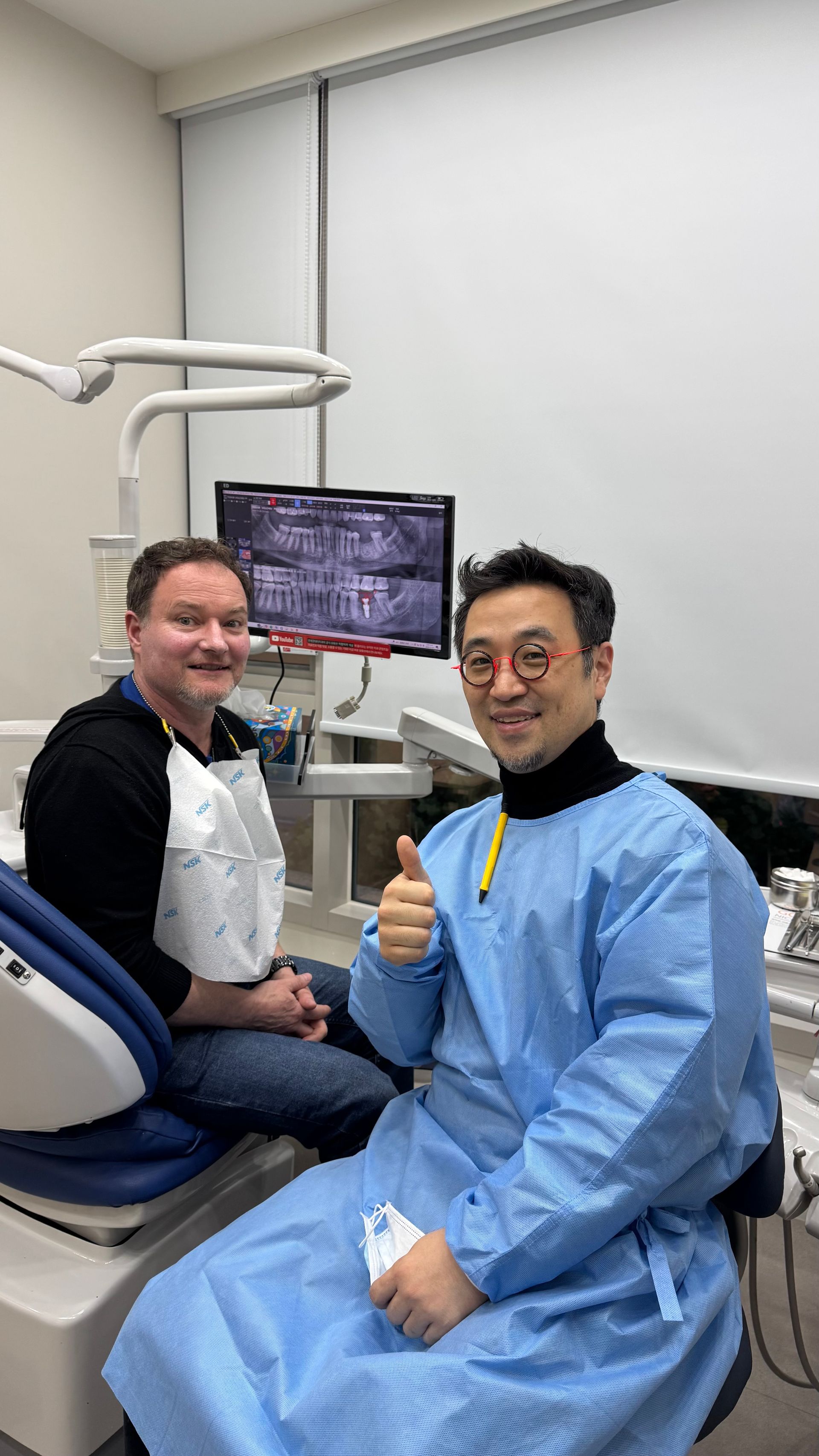 A dentist in a blue gown gives a thumbs-up next to a seated patient in a dental chair. A dental X-ray monitor is visible behind them.
