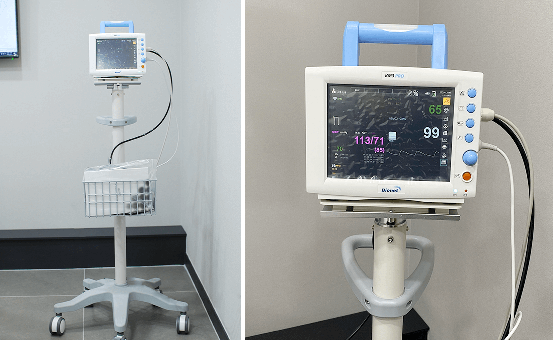 Medical monitor on a rolling stand displaying vitals. It has a blue handle and a basket for supplies.