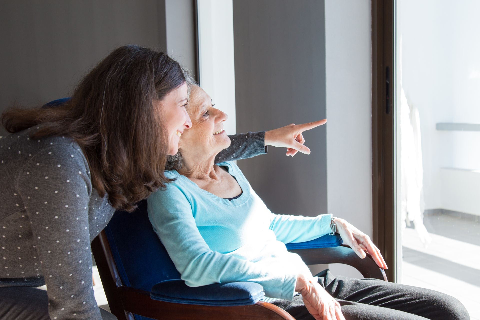 A caregiver is talking to an elderly sitting in a wheelchair while looking out the window