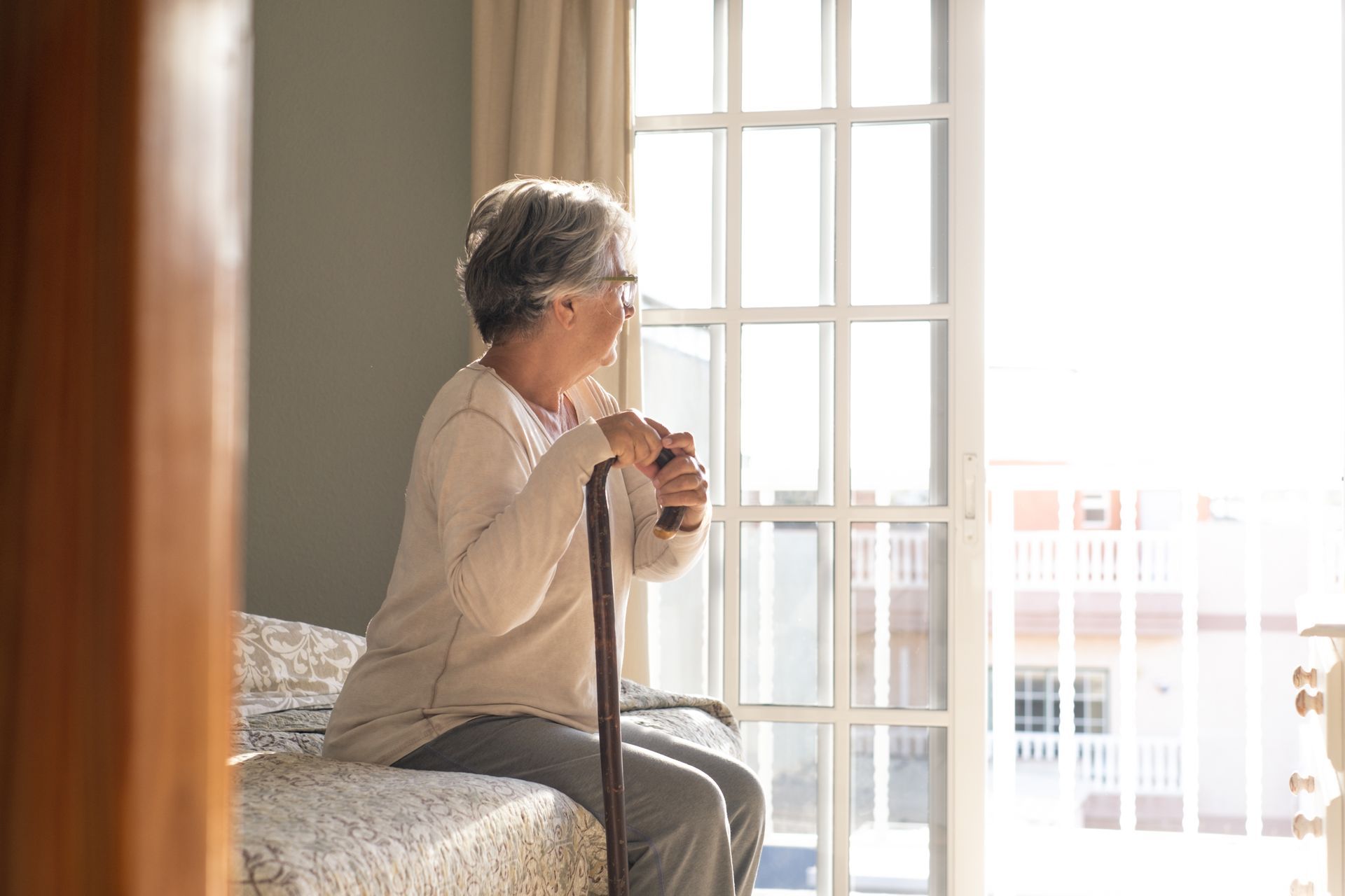 An elderly woman is sitting on a bed with a cane and looking out the window.