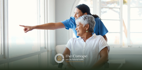 A caregiver points ahead while assisting an older adult near a window