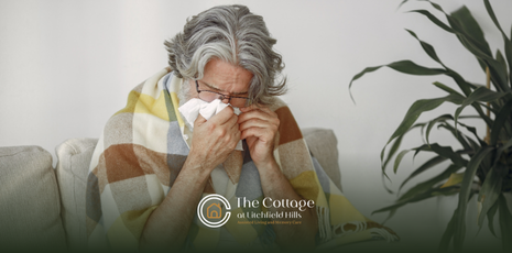 A senior sitting on a sofa wrapped in a plaid blanket, holding a tissue to his nose.