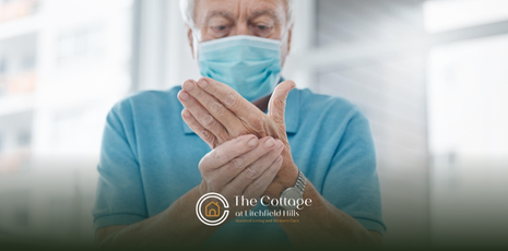 An elderly man is wearing a face mask, touching his hands with arthritis