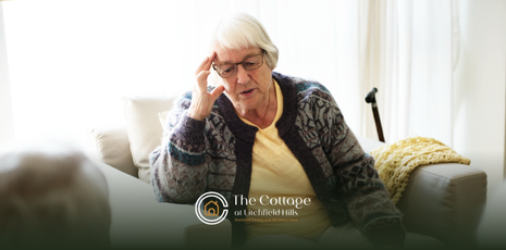 An elderly person with glasses on a couch, holding her head. 