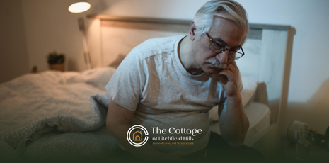 An elderly man is sitting on a bed, looking down with a hand on his face in a dimly lit bedroom.