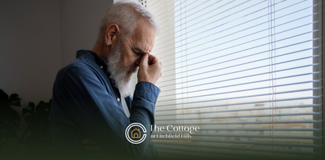 A man with a white beard, stressed, looks out a window with blinds.