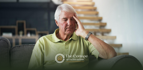 An elderly man holds his head, appearing stressed, seated on a couch. 