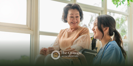 An elderly woman smiles while in a wheelchair, with a caregiver looking at her. They are near a window.