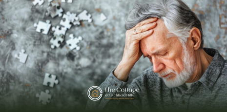 An elderly man with gray hair and beard holds his head. Puzzle pieces are scattered nearby.