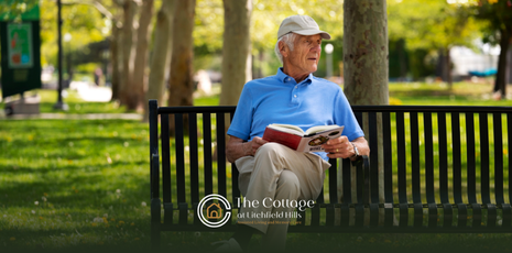 An elderly man sitting on a bench outdoors, reading a book