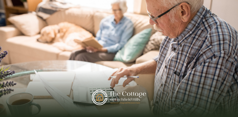 An elderly man is doing financial planning while her wife sits on a couch