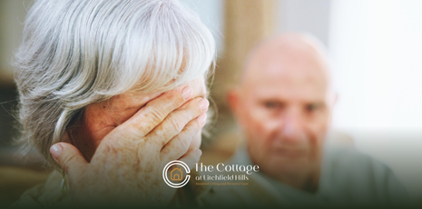 An elderly woman with her hand on her face, with her spouse behind her.