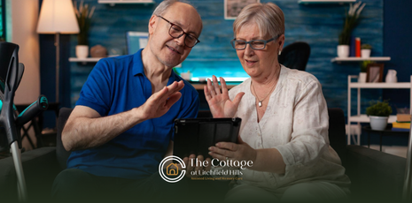 Two seniors sitting on a couch, waving at a tablet device in a senior home setting.