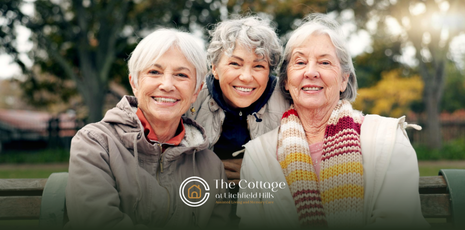 Three elderly women smiling while sitting on a park bench outdoors.