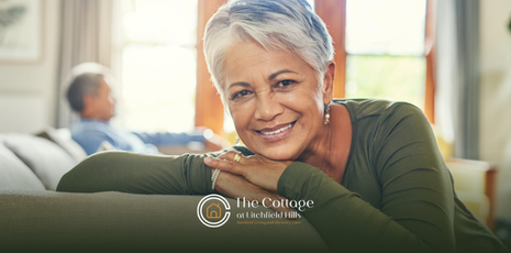 An elderly woman in her senior living apartment, smiling, resting on a couch. 