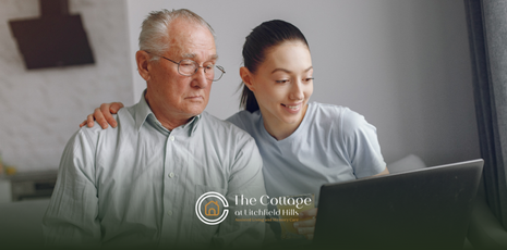 A daughter is sitting beside her senior father on a couch, looking at a laptop