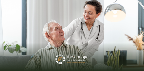 A female doctor smiles while assisting an elderly man in a living room setting.