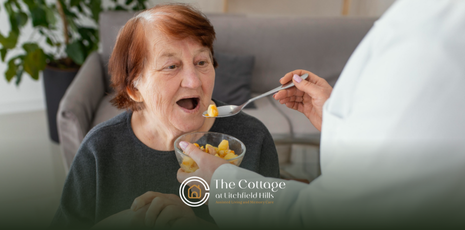 An elderly person being fed from a spoon by another person, indoors.