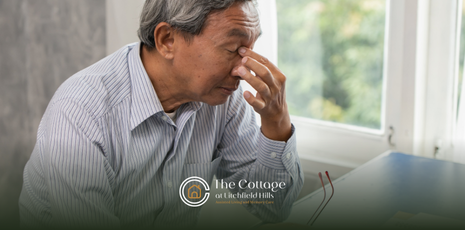 A senior sits at a table near a window, with a tired expression