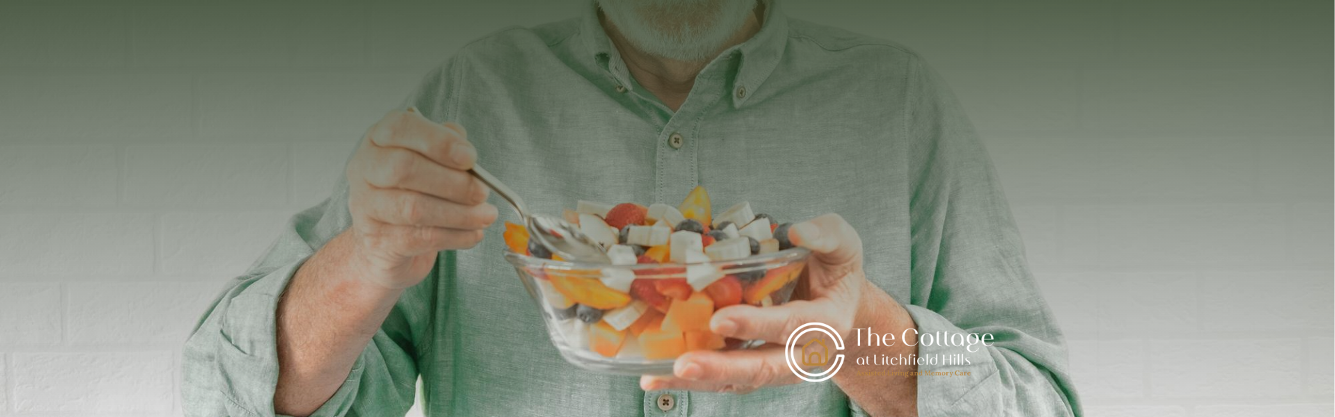 An elderly person holds a clear bowl of fruit salad, eating with a fork.