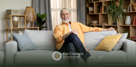 An older person with a gray beard sits on a gray couch in a living room, smiling.