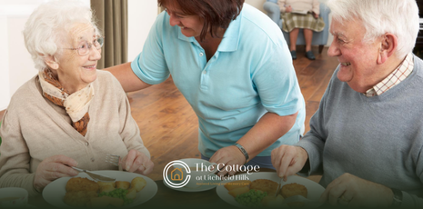 Woman assisting two seniors at a dining table. They are smiling and eating. Indoor setting.