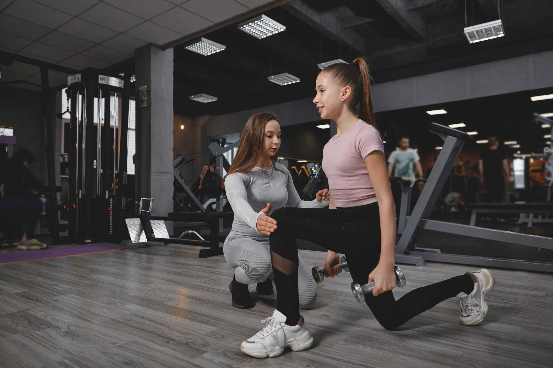 Woman guiding a girl doing a lunge with a dumbbell at a gym. Gray and pink clothing.