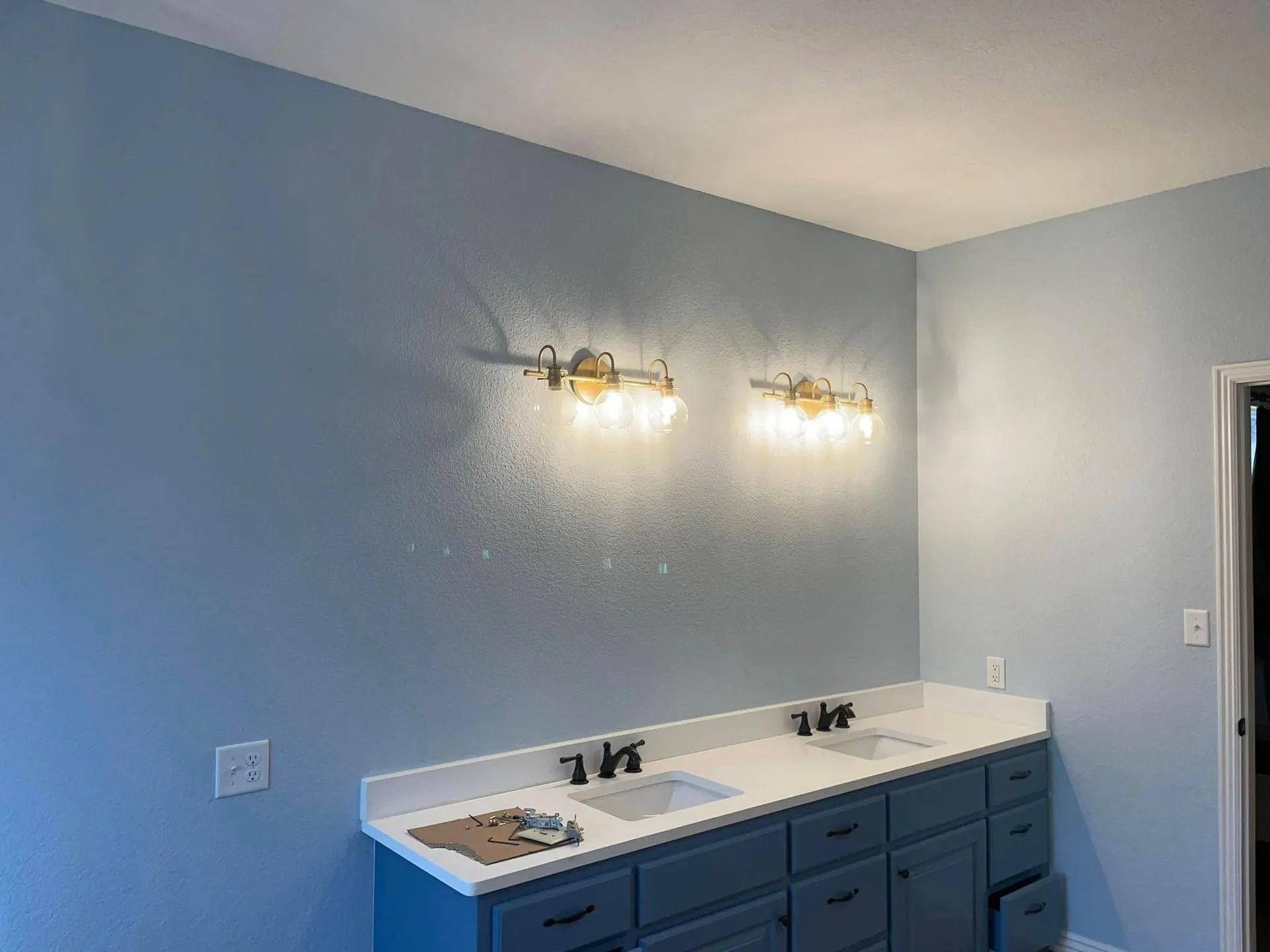 A bathroom vanity with blue cabinets, a white countertop, two sinks, and gold light fixtures against a light blue wall.
