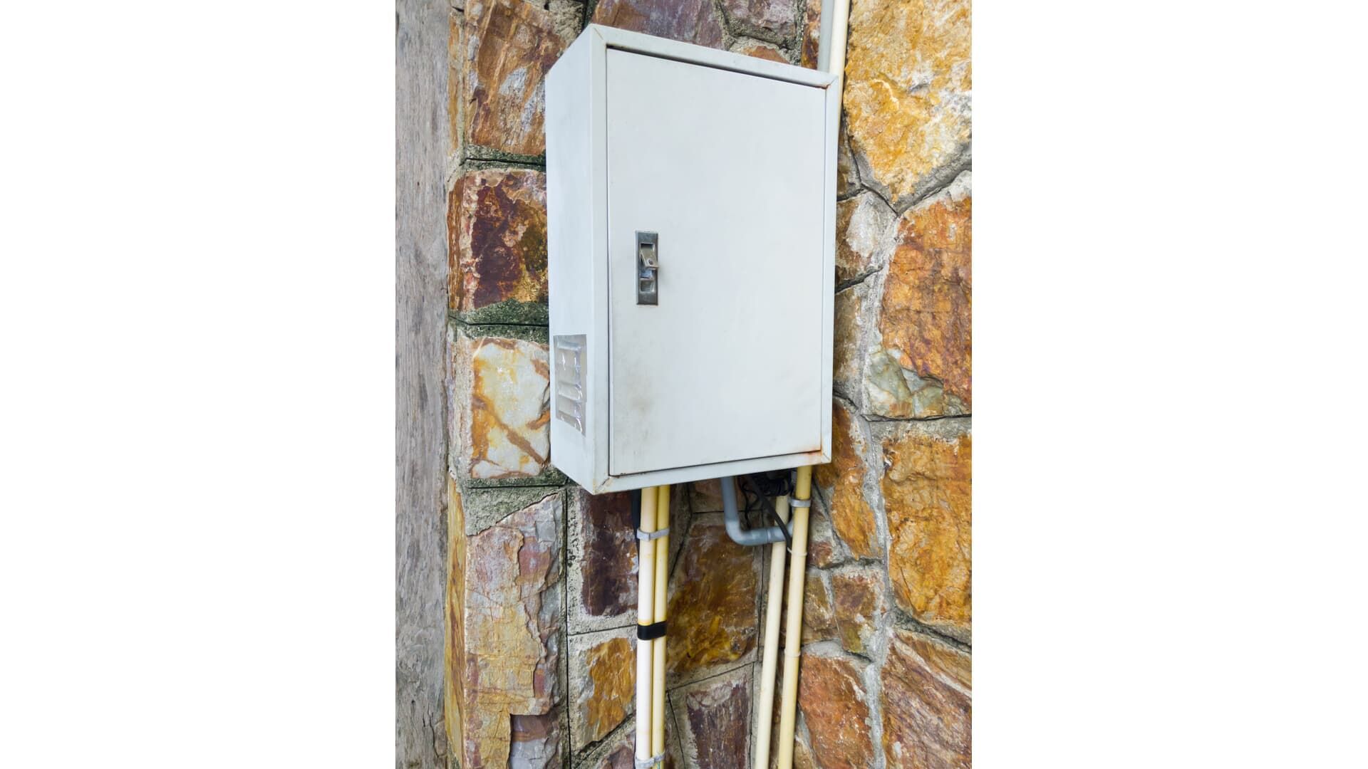 A white box is attached to a stone wall with pipes coming out of it.