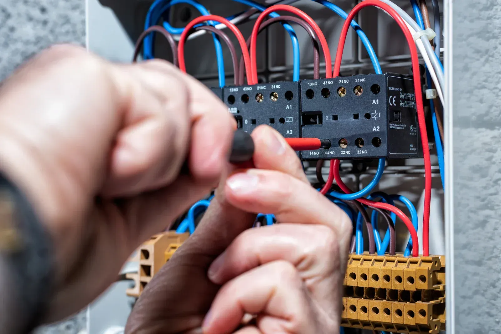 Electrician working on a panel, tightening a screw with a screwdriver. Red, blue, and white wires are visible.