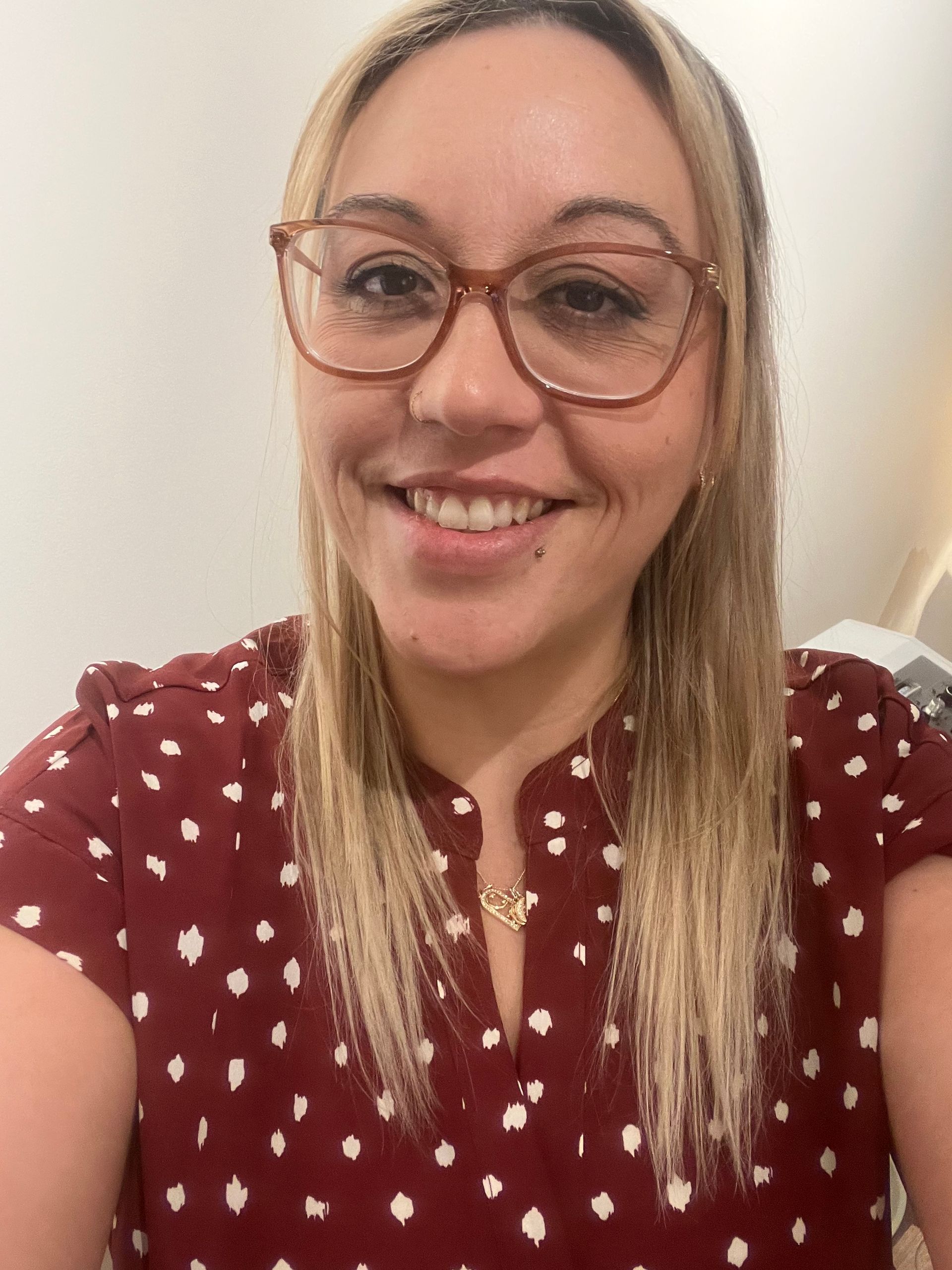 Smiling person with blonde hair and glasses, wearing a dark red patterned top, posing for a selfie.