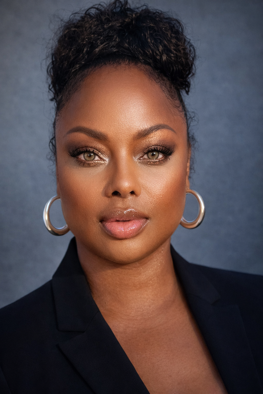 A close-up portrait of a person with curly hair pulled up, wearing silver hoop earrings and a black blazer.