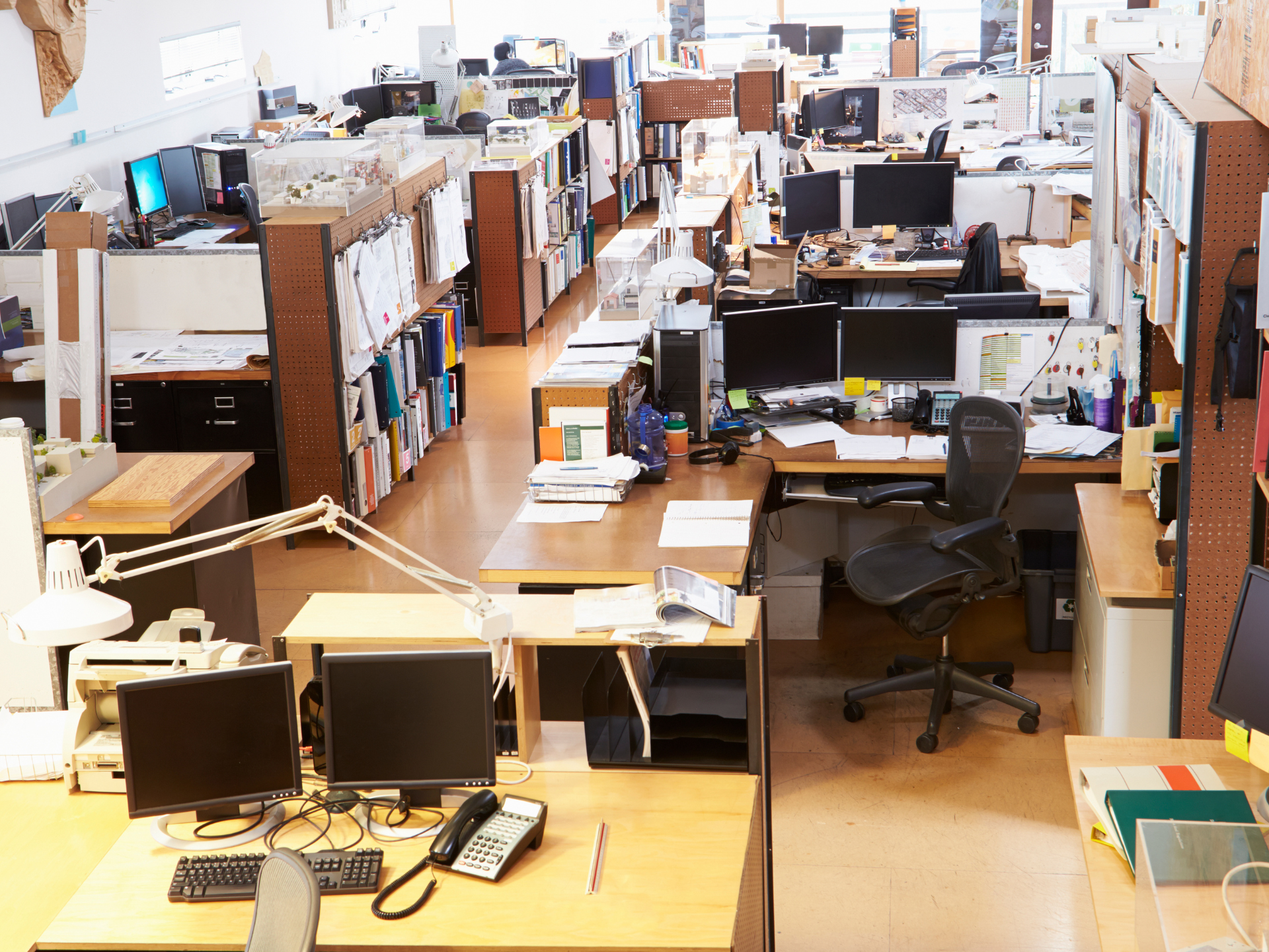 Open-plan office with desks, computers, chairs, and filing cabinets in a cluttered workspace